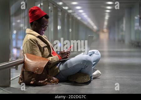 Side view of stylish African American traveler millennial man resting, sitting on the floor in empty hall of airport at night, using phone, chatting i Stock Photo