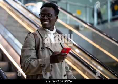 Smiling Afro-American traveler man stands on escalator in airport terminal or railway station, using mobile phone, looking aside, arrives from abroad. Stock Photo