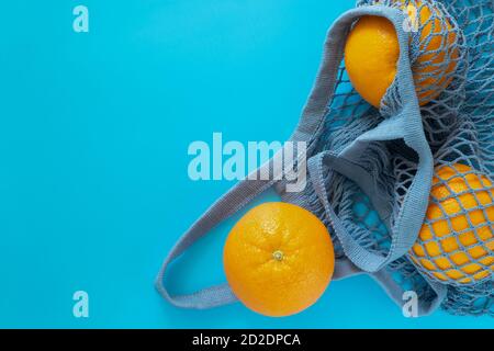 Three ripe oranges lie in a gray-blue eco-friendly string bag on a blue background. Stock Photo