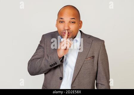 Man african american businessman in a jacket makes a gesture with his hand holding a finger on his lips, asks for silence and silence. Stock Photo