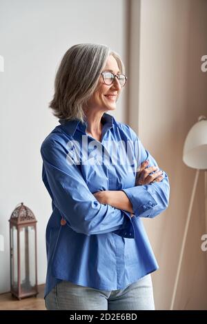 Smiling confident mature woman standing arms crossed indoors looking at window. Stock Photo