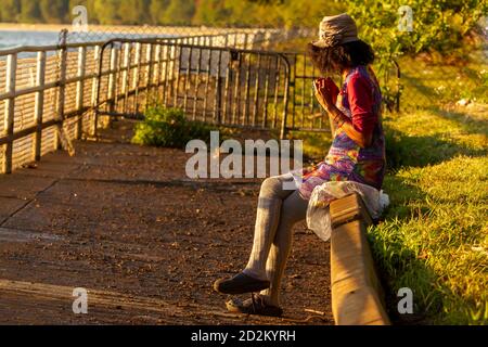 An African American woman wearing long gray socks and vibrant colored cotton dress is sitting crosslegged on a barrier wall by side walk playing with Stock Photo