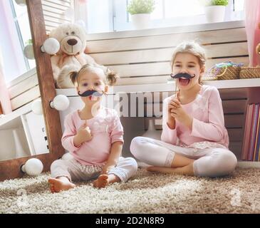 Funny family playing at home. Two cute little children girls with a paper accessories. Sisters are holding paper mustache on sticks. Stock Photo