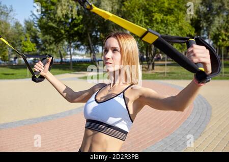 Doing TRX exercises. Young athletic woman with perfect body in sportswear training her arms with trx fitness straps in the city park Stock Photo