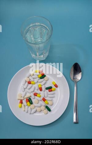 Tablets and medical capsules on white dish. Spoon and glass of clean water on blue background. The concept of pharmacology, drug overuse. Stock Photo