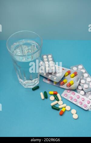 Many different pills of drugs are scattered and in blisters on a blue background. A glass of clean water for medication. The concept of treatment Stock Photo