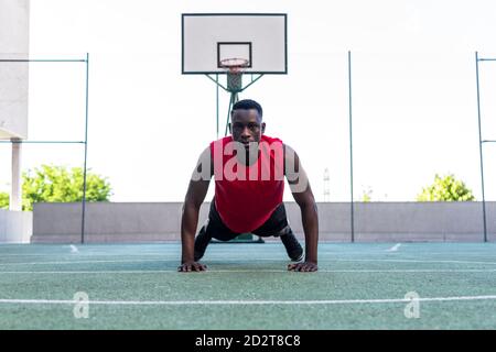 Determined black male basketball player doing push ups during intense training on sports ground and looking at camera Stock Photo