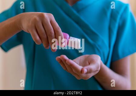 Crop of young nurse in blue scrubs and medical mask applying sanitizer on hands while preparing for procedure in clinic Stock Photo