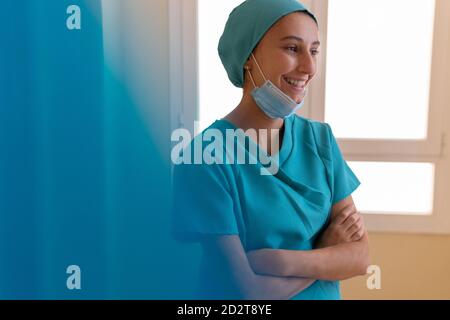 Positive young medical worker in uniform with mask standing near window and smiling happily while having break during work in hospital