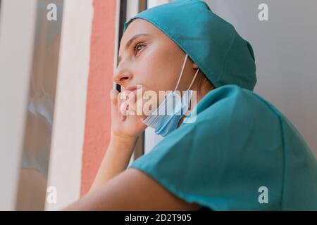 Low angle side view of thoughtful young female medical worker in uniform with protective mask on chin standing near window and looking away pensively while having rest during work in hospital Stock Photo