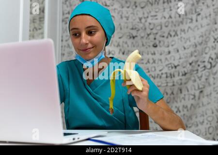 Positive young female medical worker in blue uniform using laptop and eating banana while working at nurses station in hospital Stock Photo