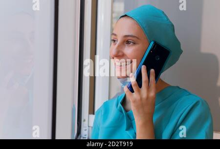 Positive young female medic in blue scrubs standing near window and chatting on mobile phone while having rest during work in hospital Stock Photo