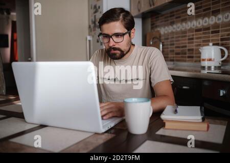 Focused young man wearing glasses using laptop, typing on keyboard, writing email or message, chatting, shopping, successful freelancer working online Stock Photo