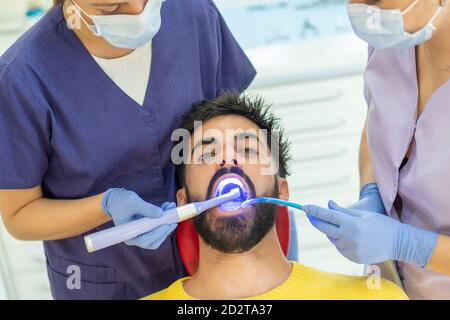 Female assistant holding suction device while dentist using mirror and drill on teeth of bearded man in clinic Stock Photo