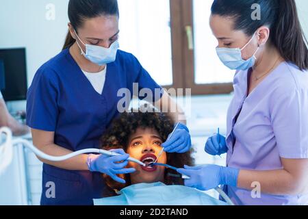 Female assistant holding suction device while dentist using mirror and drill on teeth of African American lady in clinic Stock Photo