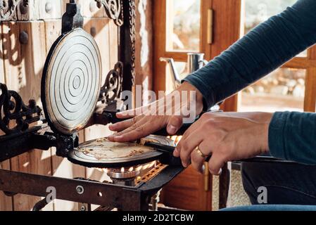 Cropped unrecognizable artisan using stick while rolling up hot freshly baked Catalan neula waffles on waffle iron placed over gas flame Stock Photo
