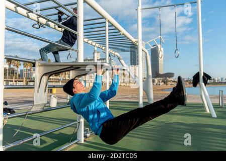 Side view of focused athletic male in activewear performing hanging leg raise exercise on metal bars during fitness workout on sports ground Stock Photo