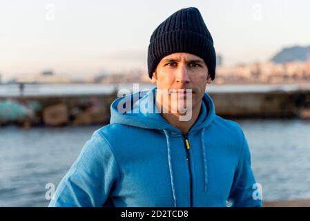 Confident adult ethnic male in warm sportive hoodie and knitted hat looking at camera while standing on embankment near sea at sunset time Stock Photo