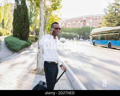 Stylish African American male entrepreneur standing with suitcase on roadside talking on the mobile phone while looking away Stock Photo