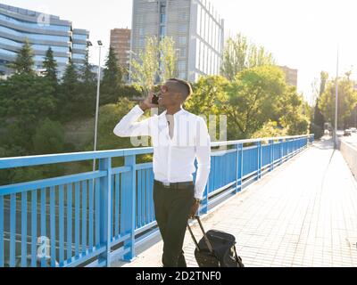 Cheerful ethnic male entrepreneur in classy wear walking with luggage along bridge and speaking on smartphone while looking away Stock Photo