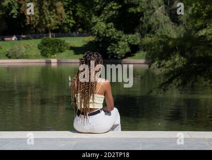 Back view of unrecognizable stylish ethnic female relaxing on embankment near pond and spending weekend in urban park in summer Stock Photo