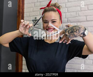 Hipster female stylist with tattoo and piercing holding scissors and looking at camera with smile while working in salon Stock Photo