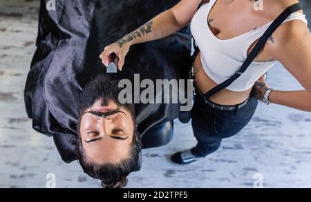 From above of crop unrecognizable female barber with trimmer in tattooed hand shaving beard of stylish brutal client with mustaches in barbershop Stock Photo
