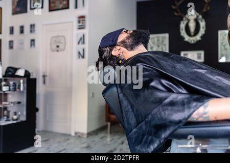 Side view of bearded hipster male client with towel on face sitting in comfortable chair and relaxing during procedure in modern barbershop Stock Photo
