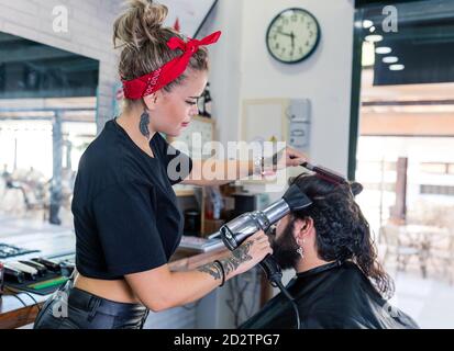 Side view of stylish female master in black leather pants drying hair of bearded male client while working in barbershop Stock Photo