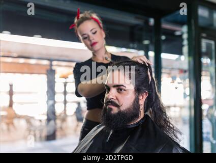 Side view of professional hipster tattooed female master setting long hair of stylish bearded man while working in modern barbershop Stock Photo