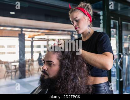 Side view of professional hipster tattooed female master setting long hair of stylish bearded man while working in modern barbershop Stock Photo
