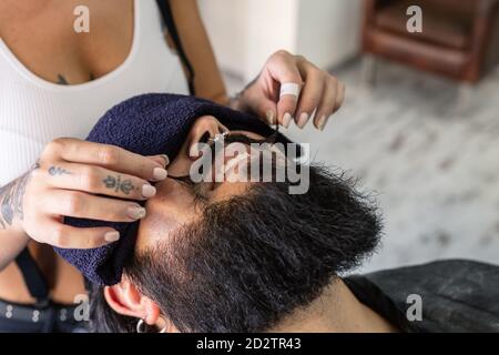 High angle of crop unrecognizable female master with tattooed body styling mustache of happy brutal bearded man with piercing during haircut procedure in barbershop Stock Photo