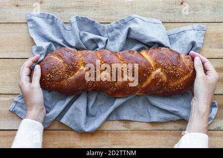 Top view of cropped unrecognizable person hands holding appetizing aromatic braided bread with seeds placed on linen napkin on wooden table Stock Photo