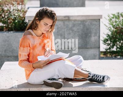 Low angle of young Woman in casual clothes sitting cross legged on border and opening notepad while doing homework outside university building in Madrid, Spain Stock Photo