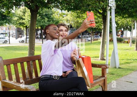 Happy stylish young multiracial female students in similar clothes sitting on bench with shopping bags and taking selfie on smartphone while chilling together in park after shopping time Stock Photo