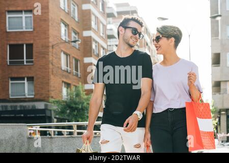 Cheerful young man and Woman in casual outfits and trendy sunglasses carrying shopping bags while walking and chatting together on street after shopping Stock Photo
