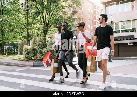 Full length of cheerful young diverse students in trendy outfits and sunglasses carrying purchases in shopping bags and chatting happily while crossing road on city street Stock Photo