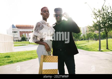 Low angle of confident young African American couple in fashionable outfits with shopping packet standing on urban square and looking at camera Stock Photo
