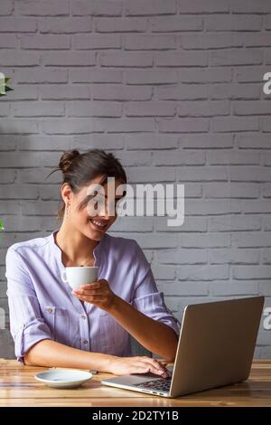 Smiling young female remote employee with cup of coffee in hand sitting at table and browsing laptop while working online from home Stock Photo