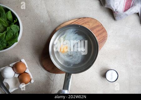 Top view of metal saucepan with boiling water and egg during poached eggs preparation on kitchen table with ingredients Stock Photo