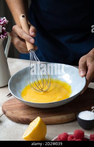 Crop unrecognizable male cook in apron standing at table with ingredients and mixing eggs with whisk in bowl while preparing breakfast in kitchen Stock Photo