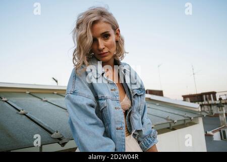 Tranquil female in denim jacket and with wavy hair relaxing in house roof in the city in evening and looking at camera Stock Photo