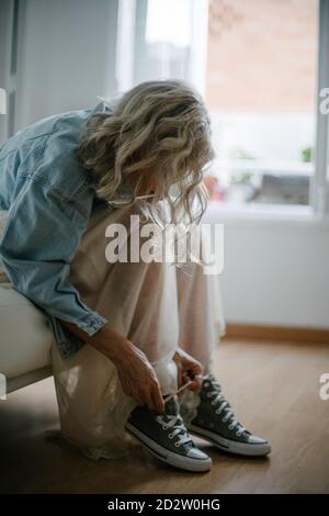 Side view of stylish female in denim jacket and skirt sitting on sofa and tying shoelaces on gumshoes Stock Photo