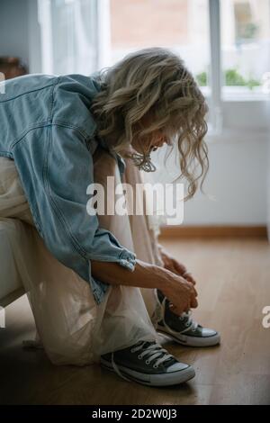 Side view of stylish female in denim jacket and skirt sitting on sofa and tying shoelaces on gumshoes Stock Photo