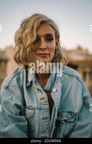 Tranquil female in denim jacket and with wavy hair relaxing in city in evening and looking at camera Stock Photo