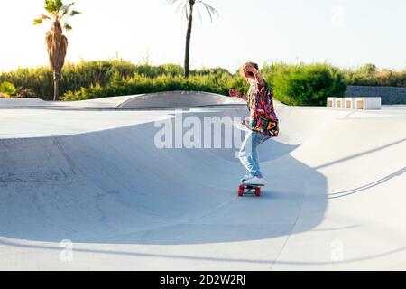 Back view of anonymous funky young male skateboarder in trendy colorful shirt and jeans performing trick on concrete ramp while practicing skills in skatepark Stock Photo