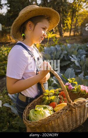 Side view of cute kid in straw hat standing in lush garden and collecting ripe vegetables in basket in summer Stock Photo