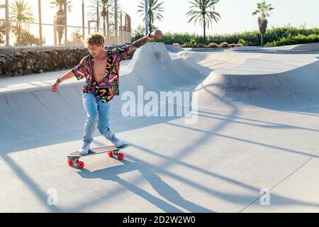 Full body of funky young male skateboarder in trendy colorful shirt and jeans performing trick on concrete ramp while practicing skills in skatepark Stock Photo