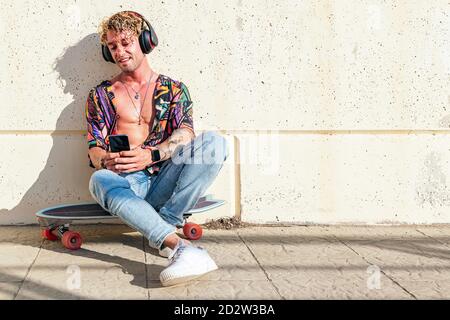 Young man hipster sitting on skateboard on street and enjoying songs in headphones Stock Photo