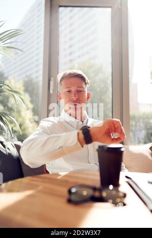 Content businessman sitting at table in modern workplace and checking time on wristwatch while waiting for meeting Stock Photo